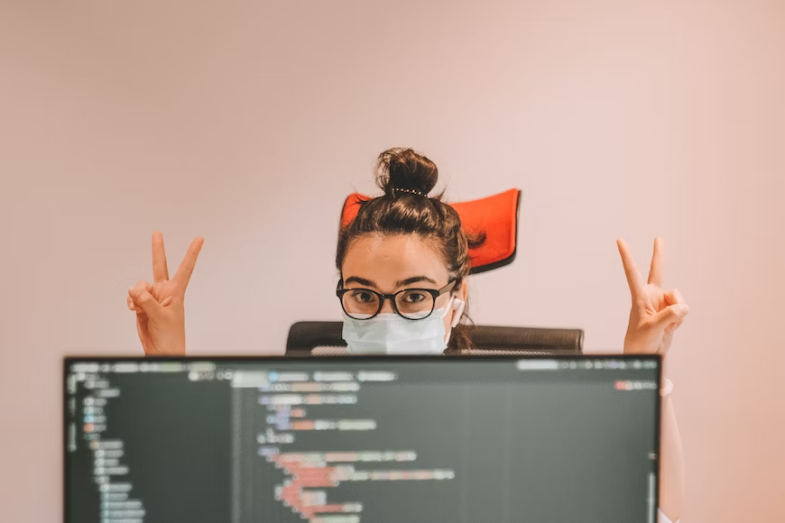 A woman, wearing a face mask, sits at a computer and raises two fingers in a gesture, indicating a sign of victory or peace.png